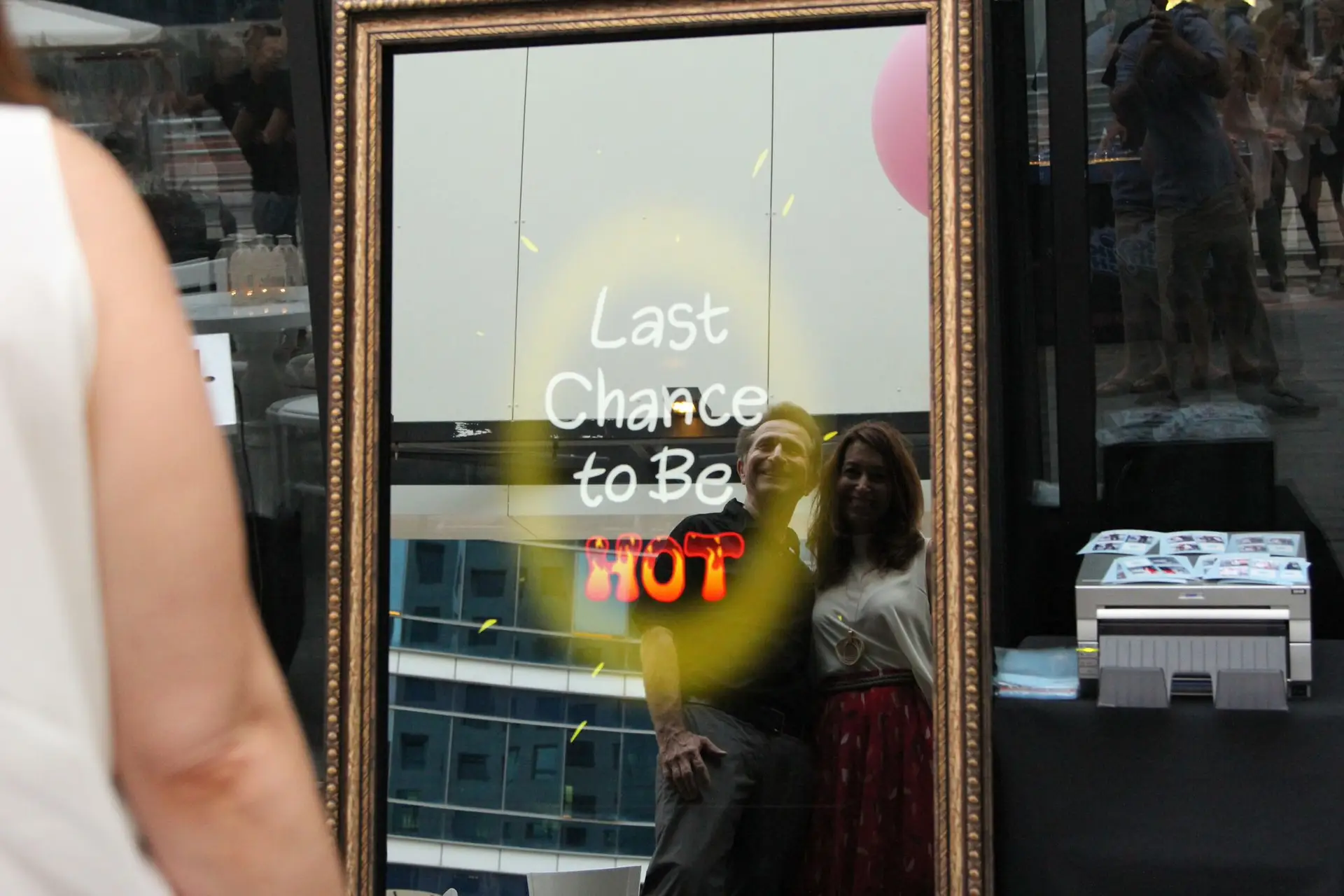 A reflection of a couple posing in front of the Parti-Panorama Photo Booth with the playful words "Last Chance to Be Hot" displayed on the mirror