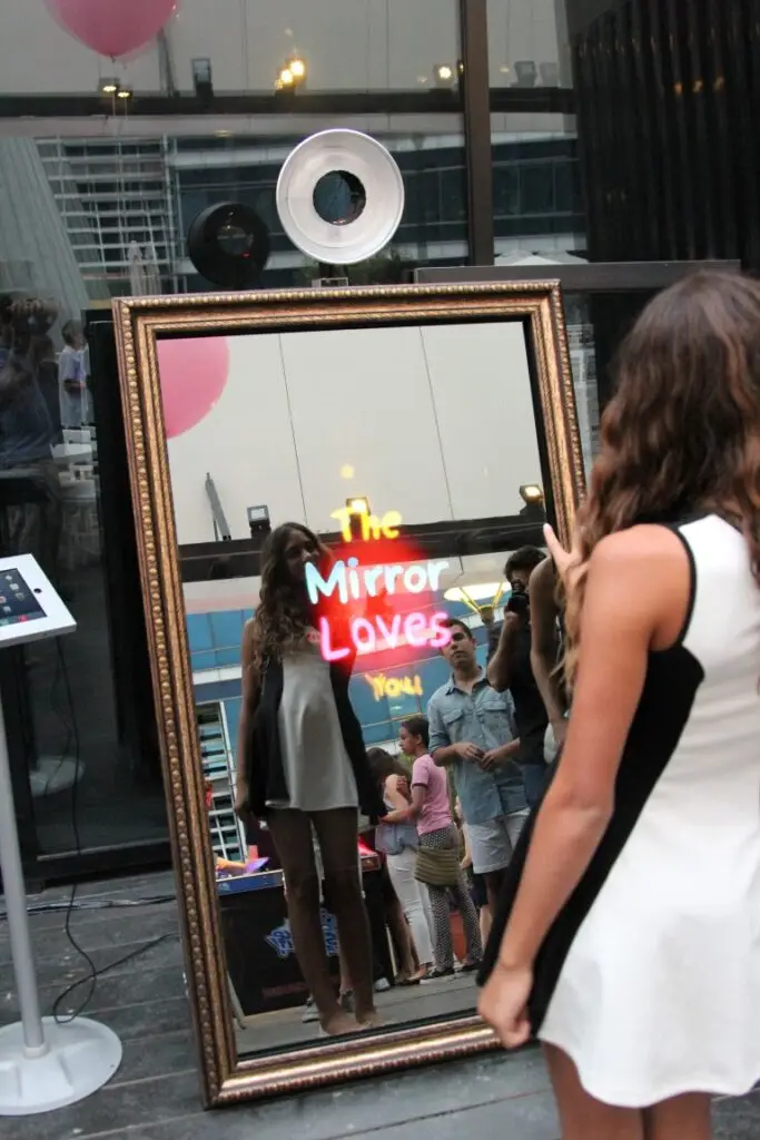 A reflection of a young woman posing in front of the Parti-Panorama Photo Booth with the playful words "The Mirror Loves You" displayed on the mirror at a family event, with other people lined up behind her