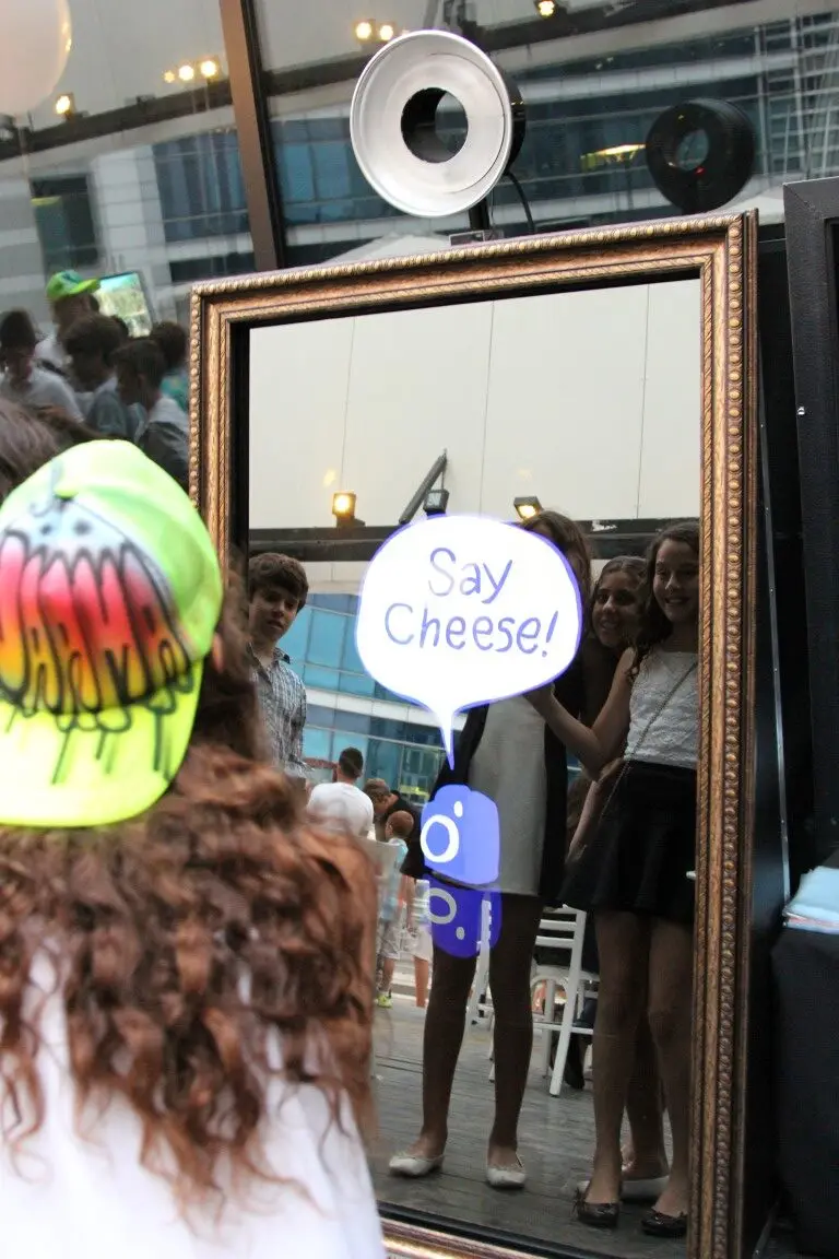 A reflection of a group of young girls posing in front of the Parti-Panorama Photo Booth with the playful words "Say Cheese" displayed on the mirror at a family event