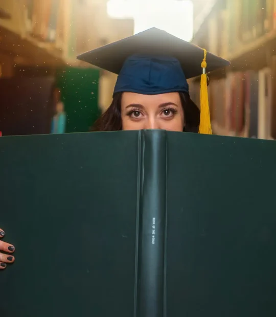 A girl wearing her graduation cap holding a book titled "Book of the World" on the spine and hiding her face behind it