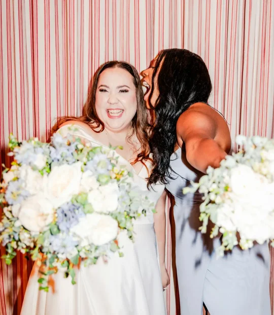 A bride and her bridesmaid posing cheekily together in front of a red and white stripe backdrop