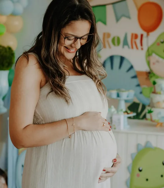Smiling pregnant woman at a dinosaur themed baby shower, holding her belly.