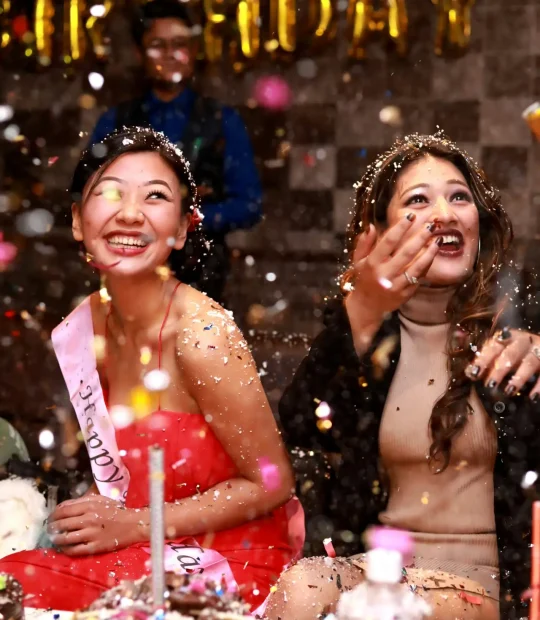 Two women, one wearing a birthday sash, admiring the confetti falling down at a birthday party
